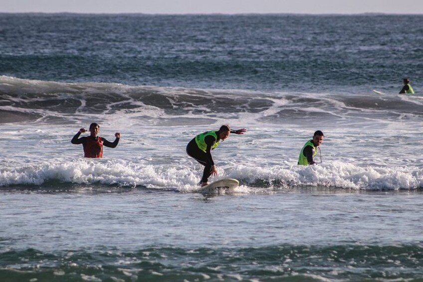 Three surfers in the ocean, one in a red shirt and two in green shirts, with waves and a clear sky in the background. 4