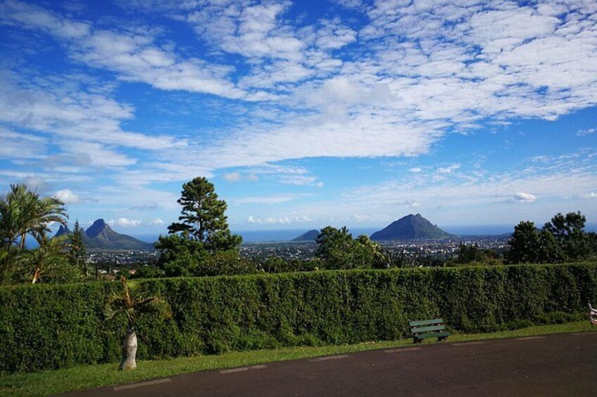 A road with a bench, a hedge, and a view of mountains and a city in the distance. 2