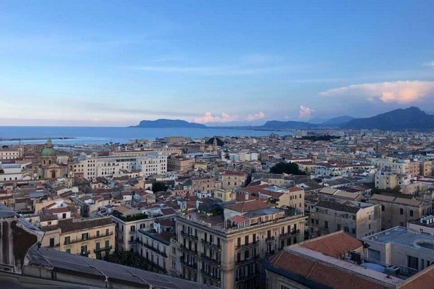 A coastal cityscape with historic buildings, a clear sky, and distant mountains. 1