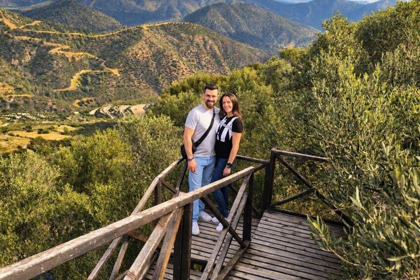 Eine Holzbrücke, auf der ein Mann und eine Frau stehen und einen Blick auf eine gebirgige Landschaft mit grünen Flächen und gewundenen Pfaden werfen. 4