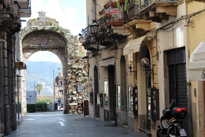 A narrow street with an old archway, buildings with balconies, and a parked scooter. 3