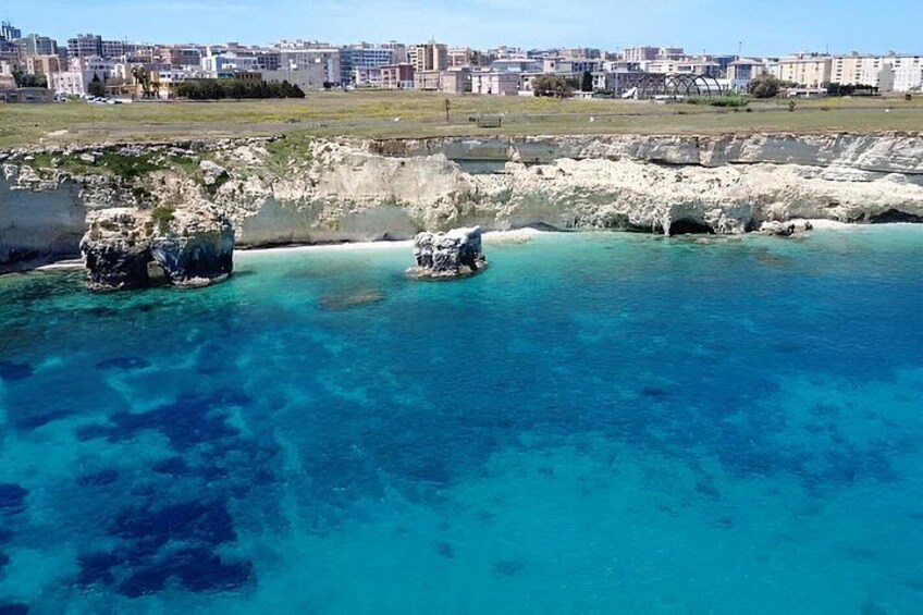 A coastal area with clear blue water, rocky formations, and a distant cityscape. 4