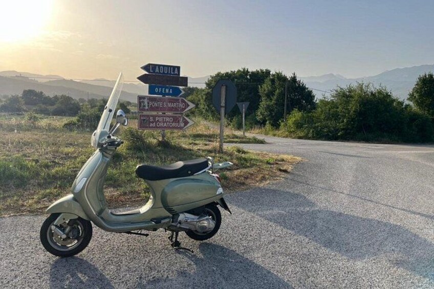 A vintage Vespa scooter parked on a rural road with directional signs pointing to various destinations. 1