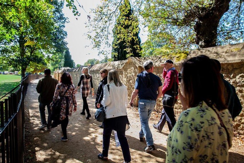 Un grupo de personas caminando por un sendero de tierra en un parque, con árboles y un muro de piedra al fondo. 5