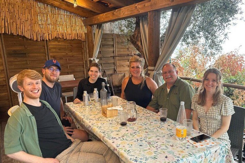 Un groupe de personnes est assis autour d’une table à l’extérieur, recouverte d’une nappe à motifs floraux, et est entouré de structures en bois et d’arbres. 4