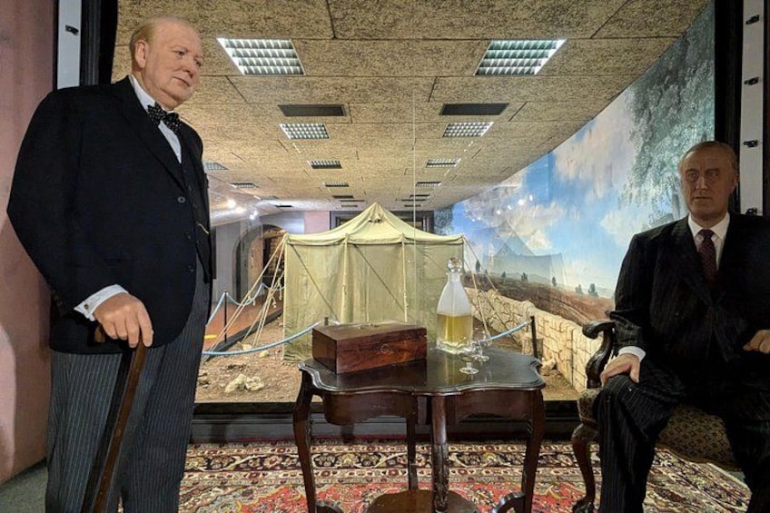 Dos hombres en trajes, uno de pie y otro sentado, en una habitación con un mural grande, una mesa con una botella y una estructura parecida a una carpa. 5
