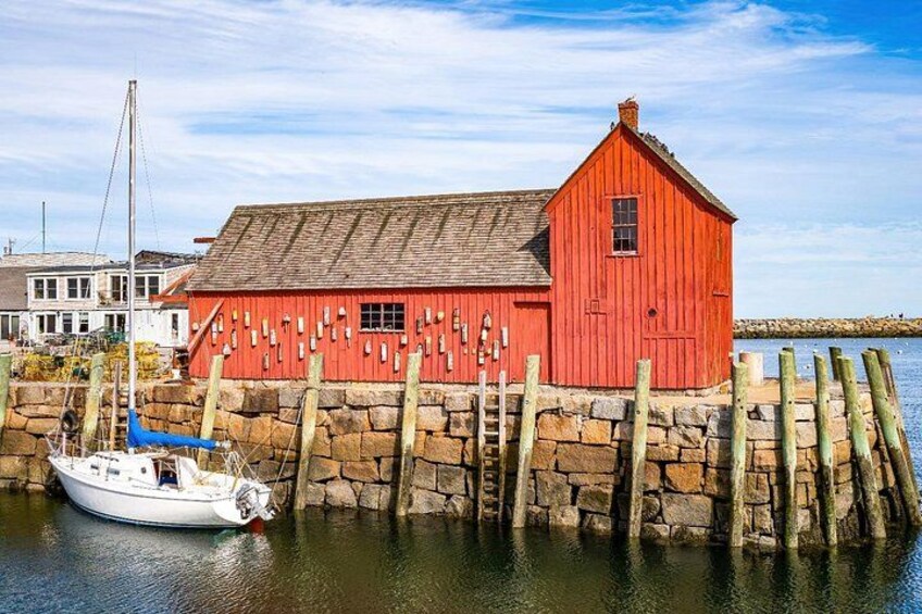 Een rood houten gebouw met een stenen steiger die uitloopt in kalm water, een witte boot afgemeerd aan de steiger, en een helder blauwe lucht. 4