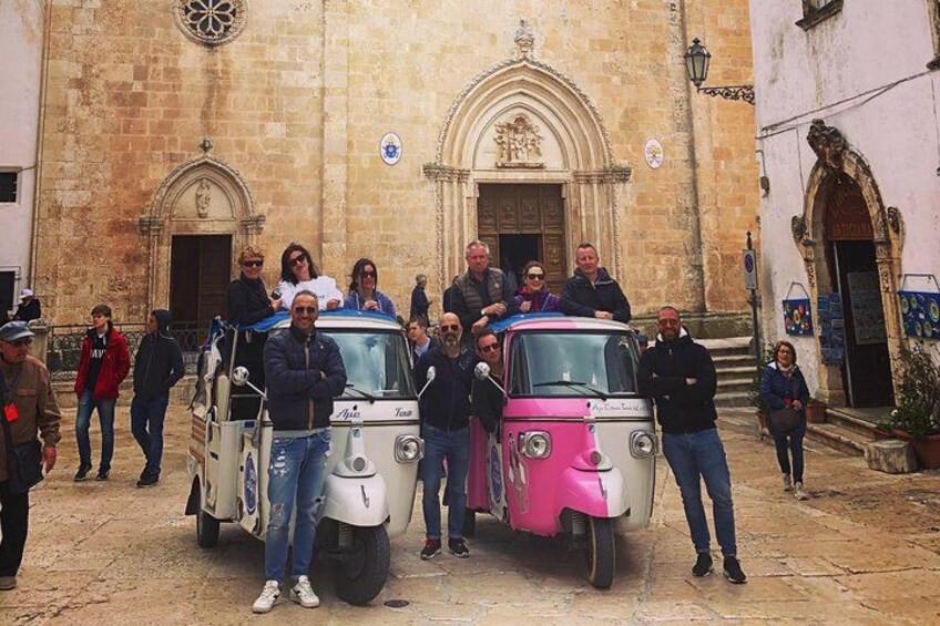 A group of people standing in front of a historic building with two vintage cars parked in front. 3