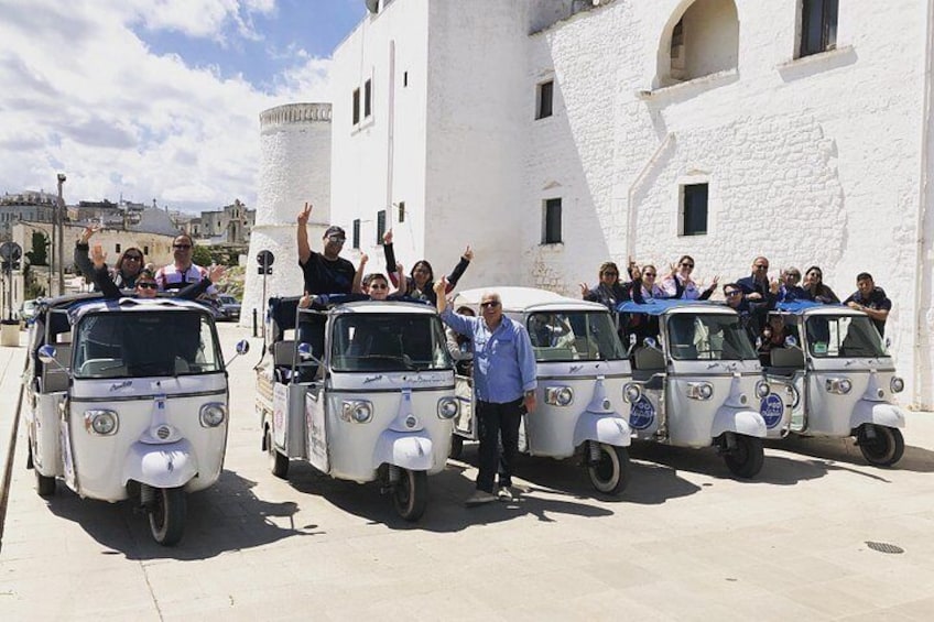 Un grupo de personas en pequeños vehículos blancos de tres ruedas frente a un edificio blanco con una torre. 1