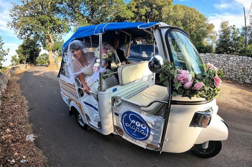 Un vehículo decorado con temática de boda, con una novia y un novio, flores y un muro de piedra al fondo. 5