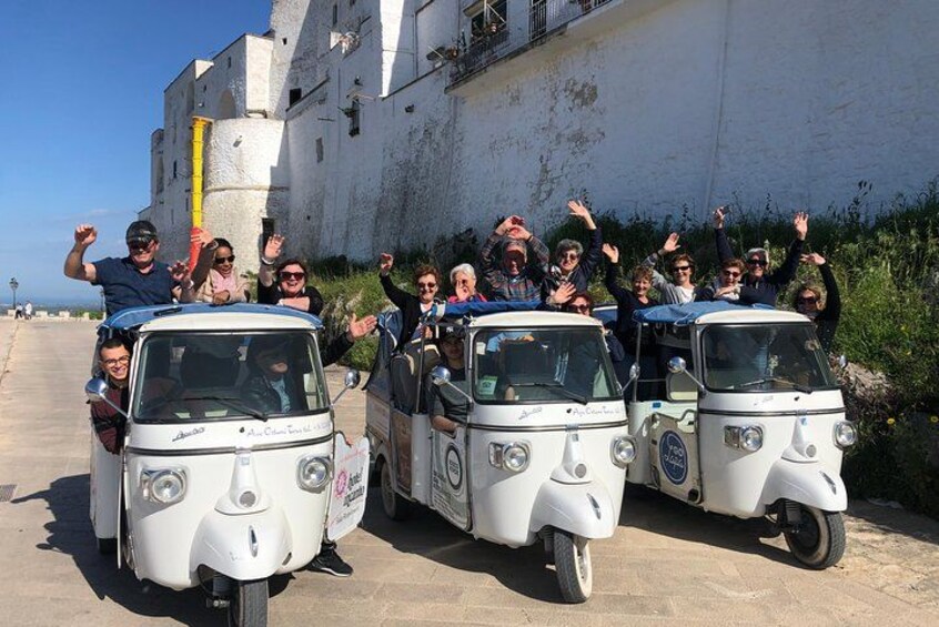 A group of people on three small white vehicles with open tops, parked on a paved area with a white building in the background. 2