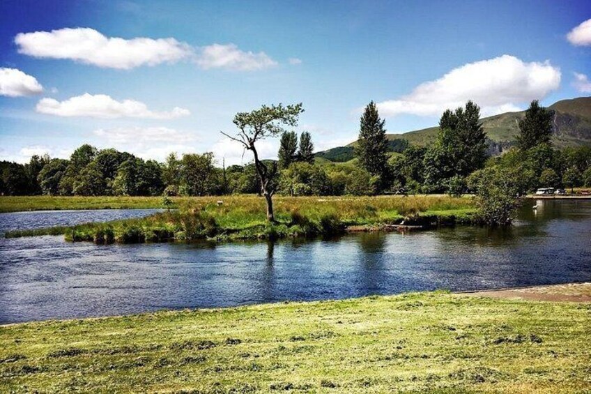 Ein Fluss, grasbewachsene Ufer, Bäume und Hügel im Hintergrund. 2