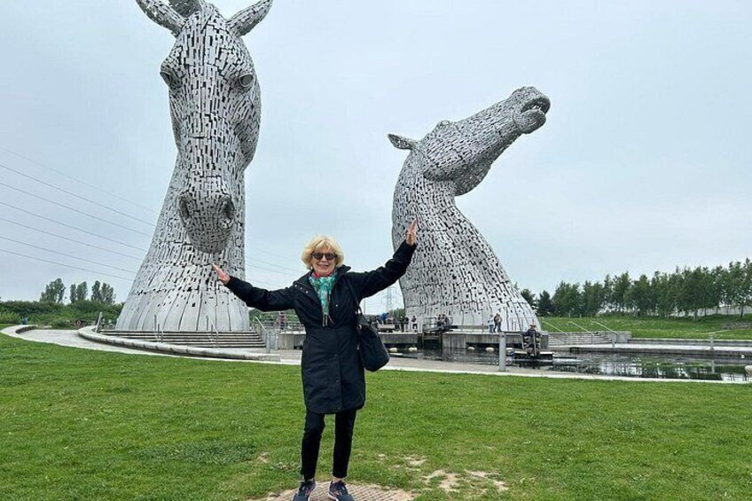 Eine Person steht vor den Kelpies, zwei großen Pferde-Skulpturen, mit einem grasbewachsen Feld und einem Gewässer im Hintergrund. 4