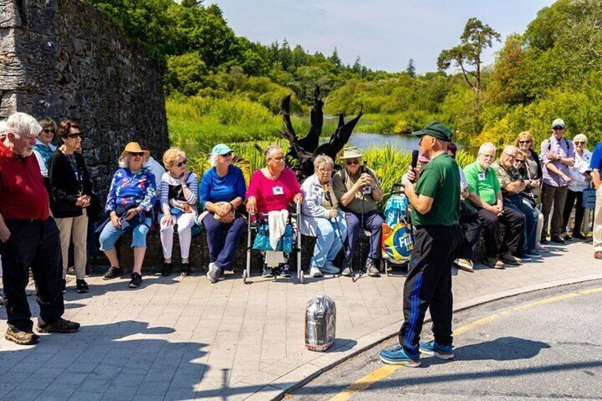 A group of people listening to a speaker outdoors with a lake and trees in the background. 3