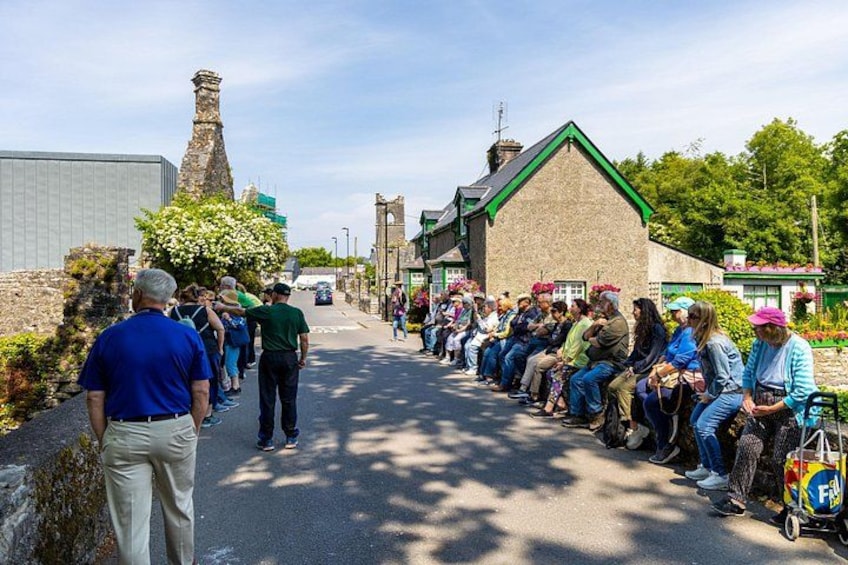 A group of people sitting on benches along a road, with buildings and greenery in the background. 2