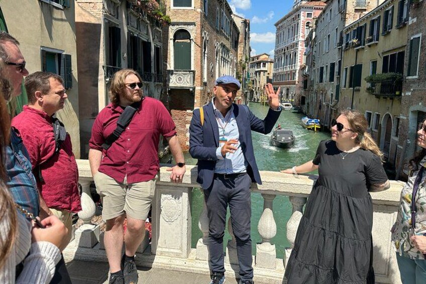 Eine Gruppe von Personen steht auf einer Brücke in einem schmalen venezianischen Kanal, flankiert von Gebäuden. 2