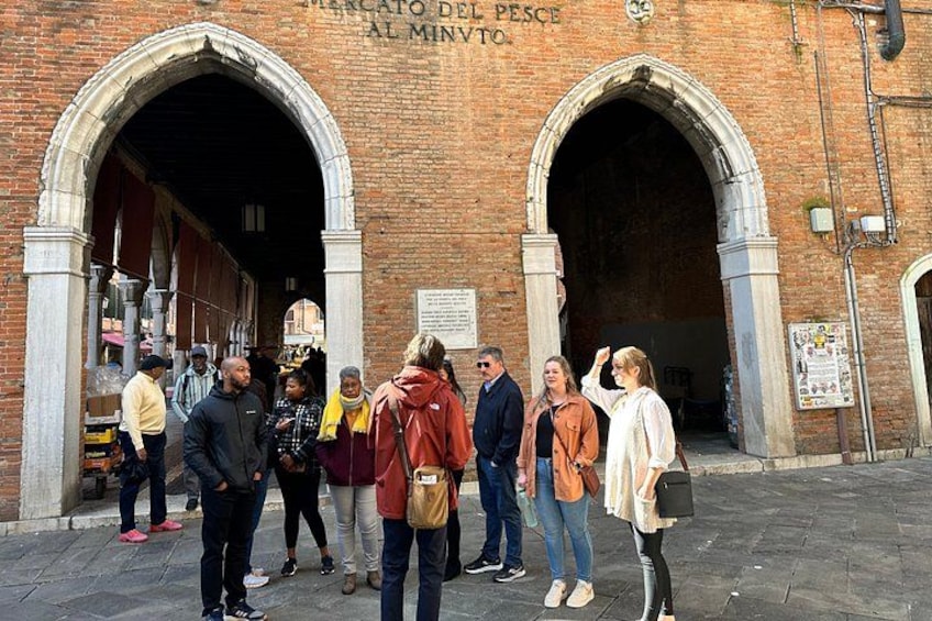 A group of people standing in front of a historic building with arched entrances, a sign reading 'Mercato del Pesce al Minvento,' and a brick facade. 3