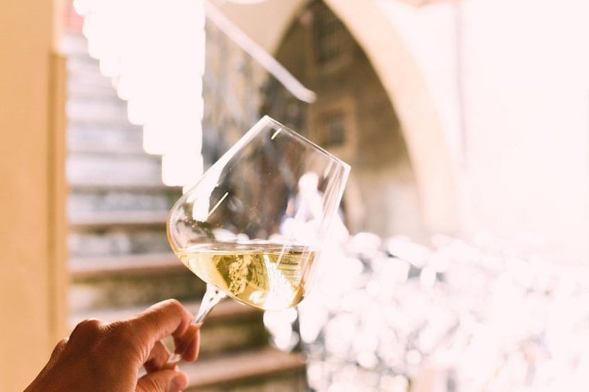 A hand holding a wine glass with a white liquid, possibly wine, in a well-lit indoor setting with architectural elements like arches and columns. 1