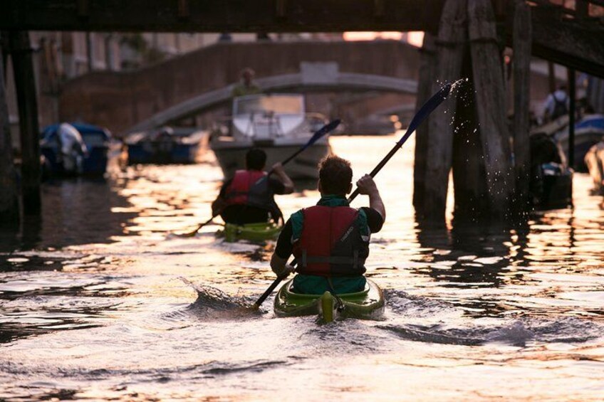 A person in a green kayak paddling under a bridge, with another person in a red kayak in the background. 2