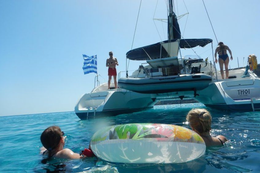 Une personne nage dans l’océan, tenant un flotteur gonflable aux couleurs vives. On aperçoit également un voilier avec des drapeaux et un chien dans l’eau. 4