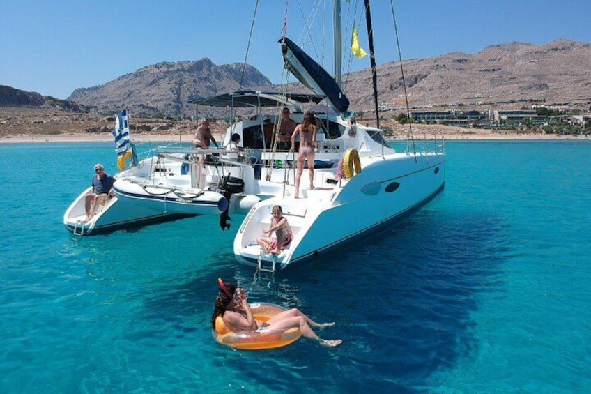 Un yacht blanc avec des personnes à bord, une femme en maillot de bain rose flottant sur un bouée gonflable orange, et un paysage montagneux en arrière-plan. 1