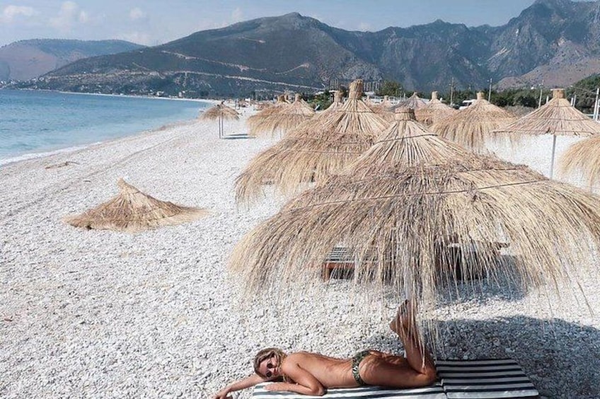 A beach with white pebbles, a person sunbathing, and thatched umbrellas. 1