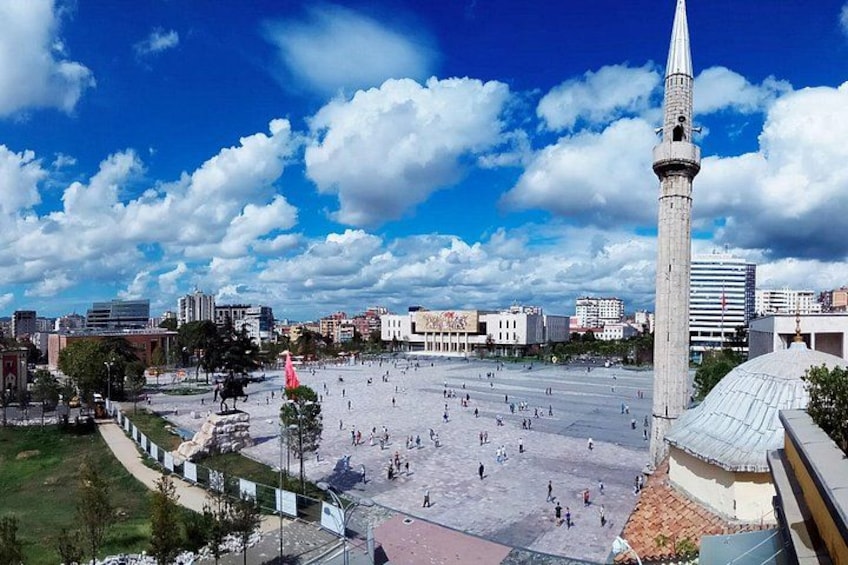 A large open square with a tall minaret, surrounded by modern buildings and a clear sky. 4