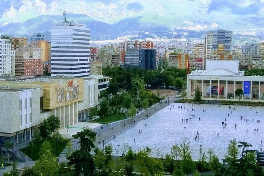 A cityscape with a large open plaza, a building with an opera house, and a mountain range in the background. 5
