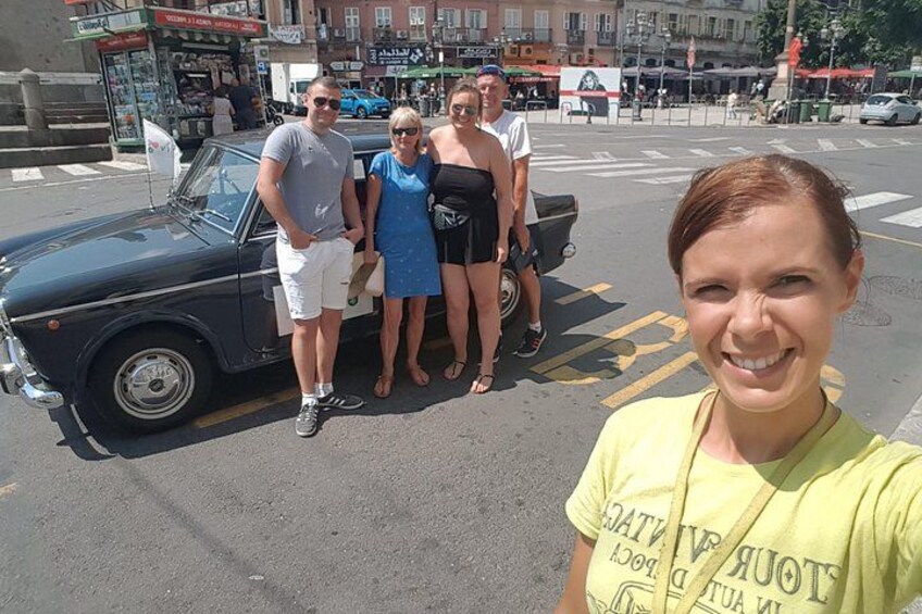 Un grupo de personas de pie junto a un coche clásico en una calle con edificios al fondo. 3