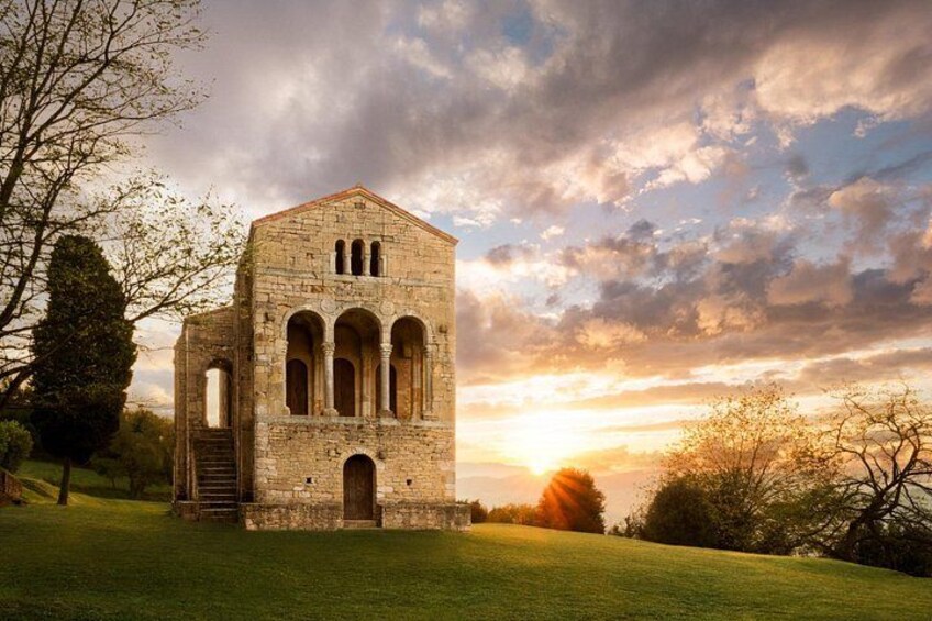 A stone building with arches, a grassy field, and a sunset in the background. 2