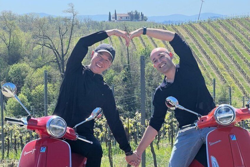Two people on red scooters in a vineyard with rows of grapevines and a distant building. 5