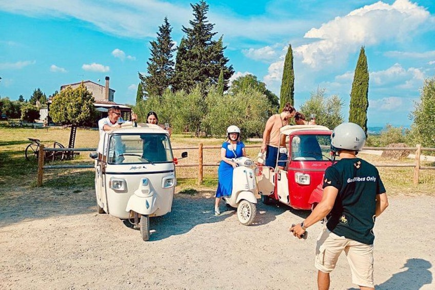 A group of people on scooters, a red vehicle, and a white vehicle in a rural setting with trees and a house in the background. 3