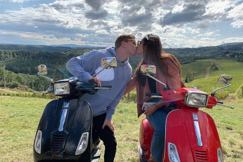 Two people on scooters, a man and a woman, kissing in a grassy field with a hilly landscape in the background. 4