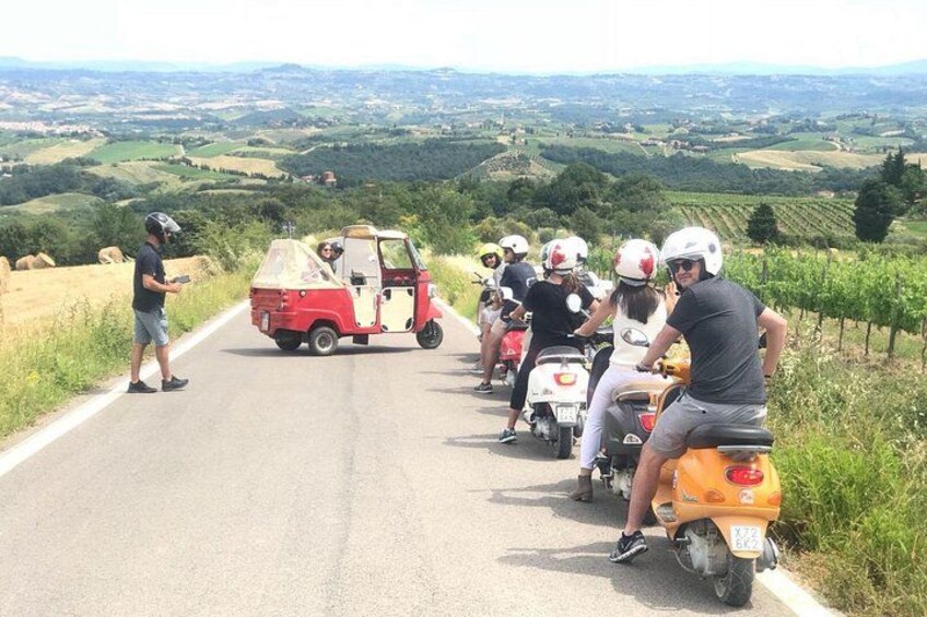 A group of people on scooters and a small vehicle on a road with a scenic landscape in the background. 3