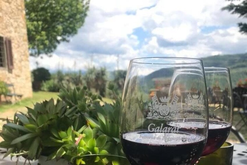 Dos copas de vino con logotipos en relieve sobre una mesa, con un edificio de piedra y follaje al fondo. 2