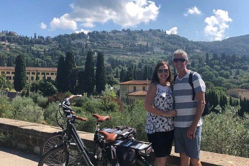 Una pareja parada junto a una bicicleta, contemplando un paisaje pintoresco con colinas y edificios. 4