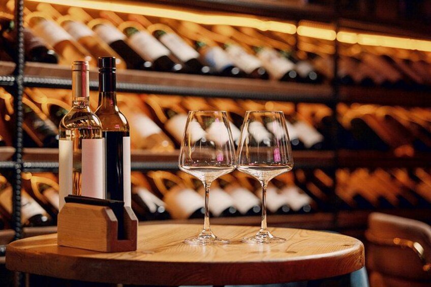 A wine bottle and two glasses on a wooden table with a wine rack in the background. 1
