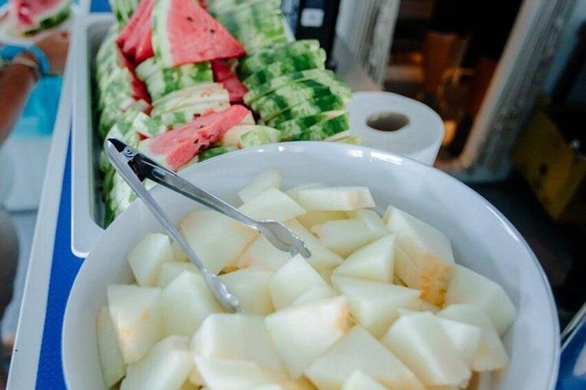 A bowl of cubed apples with a pair of tongs resting on top, and a tray of sliced watermelon in the background. 2