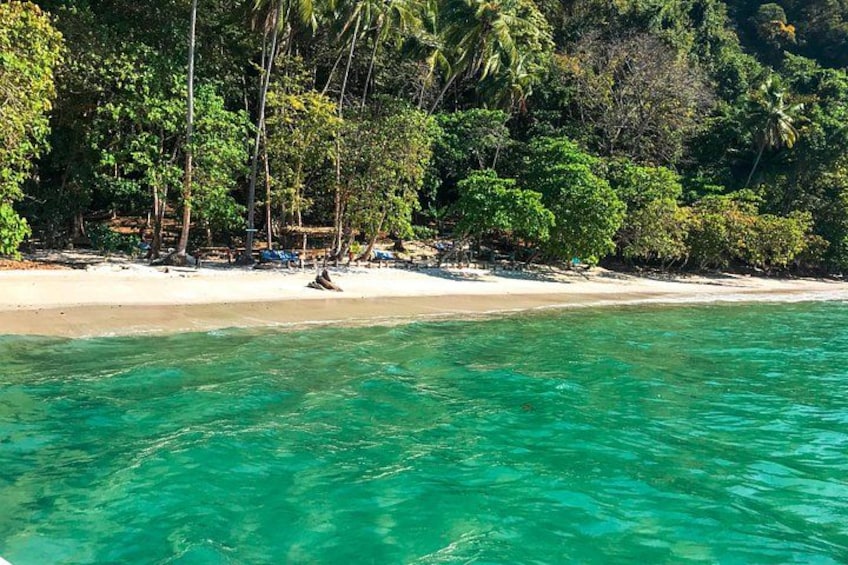 A beach with clear turquoise water, white sand, and dense tropical foliage. 1