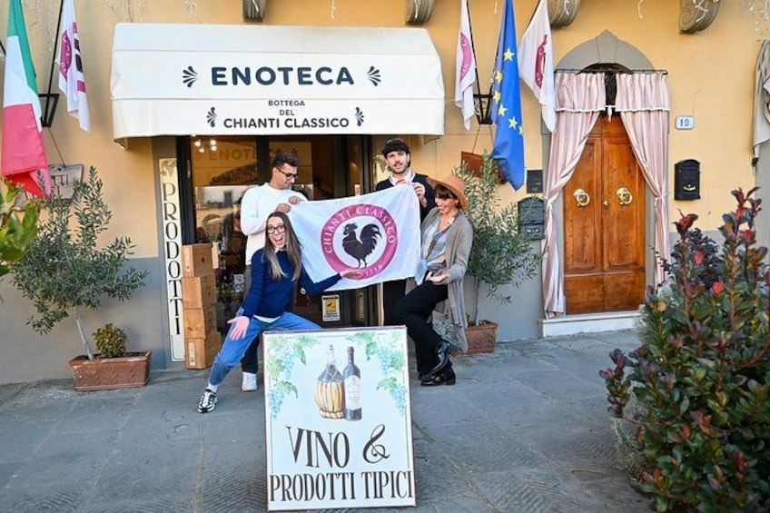 Una vetrina con un cartello che recita 'ENOTECA' e 'CHIANTI CLASSICO'. Si vedono bandiere, una porta in legno e un cartello con la scritta 'VINO & PRODOTTI TROPICI'. 5