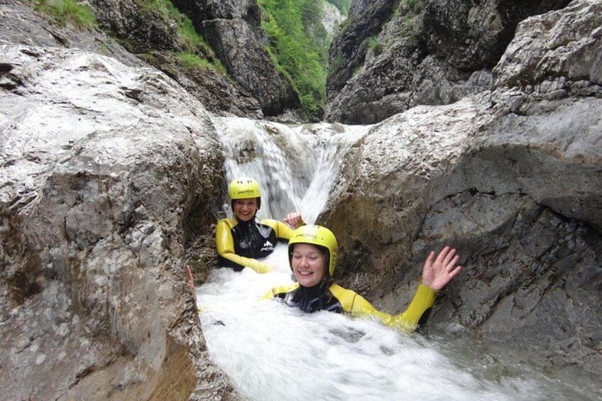 Two people in yellow helmets and wetsuits sitting in a rocky water channel with a waterfall above them. 1