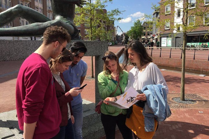 Un groupe de personnes se tient sur une place pavée de briques, avec une grande sculpture en arrière-plan. 3
