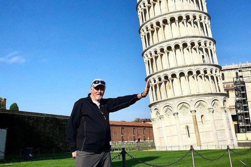 Una persona de pie junto a la Torre Inclinada de Pisa, con una vista clara de la inclinación particular de la torre y el área verde circundante. 1