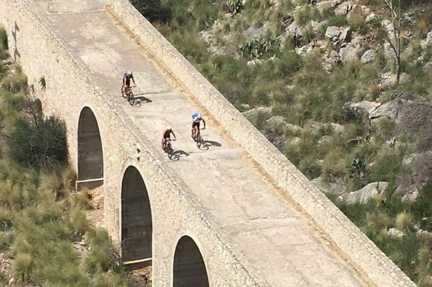 En stenbro med valv, cyklister som cyklar över den och ett bergigt landskap med gles vegetation. 5