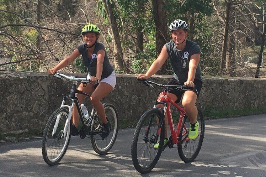 Zwei Radfahrer auf einer asphaltierten Straße, im Hintergrund eine Steinmauer und Bäume. 4