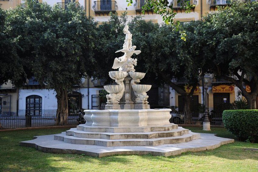 A stone fountain with multiple tiers and water spouts, surrounded by grass and trees, with buildings in the background. 5