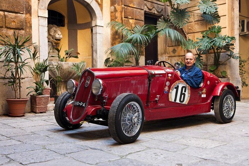 Un coche de carreras rojo antiguo con el número 16 estacionado frente a un edificio con arcos y plantas en macetas. 2