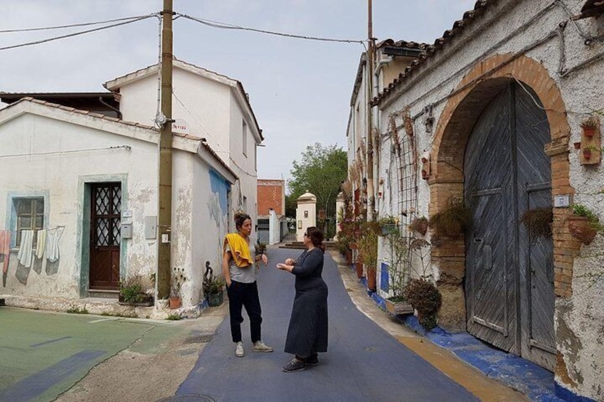 A narrow street with buildings on either side, a woman in a long dress, and a man in a yellow shirt. 1