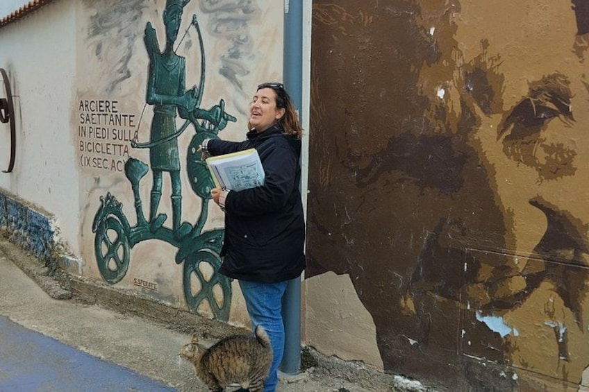 A person leaning against a wall with a mural of a figure on a bicycle, holding a book. 4