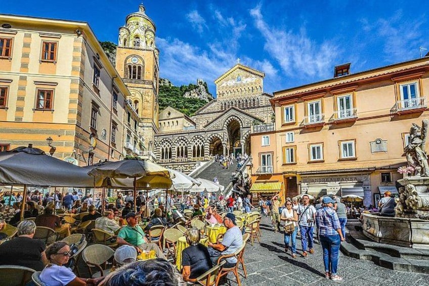 A crowded square with people sitting at tables, a fountain, and historic buildings. 2